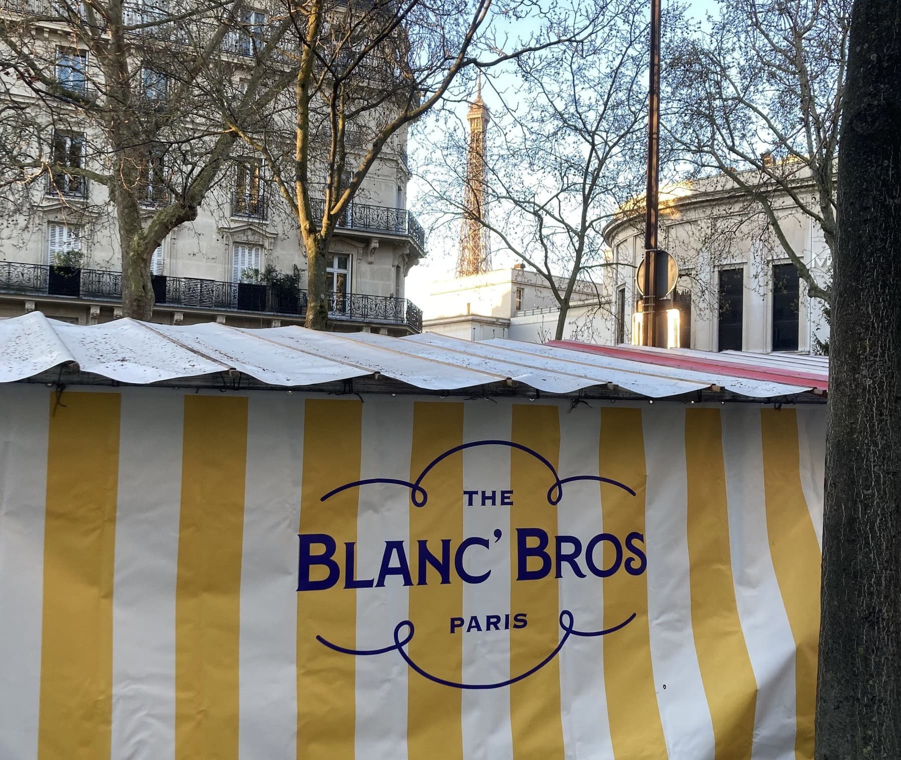 Étal du marché The Blanc' Bros à Paris, banderole rayée jaune et blanche avec vue sur la Tour Eiffel